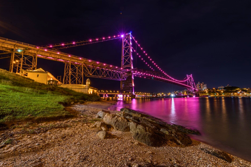 Ponte Hercílio Luz iluminada em rosa à noite, com reflexo nas águas da baía de Florianópolis. Custo de vida em Florianópolis
